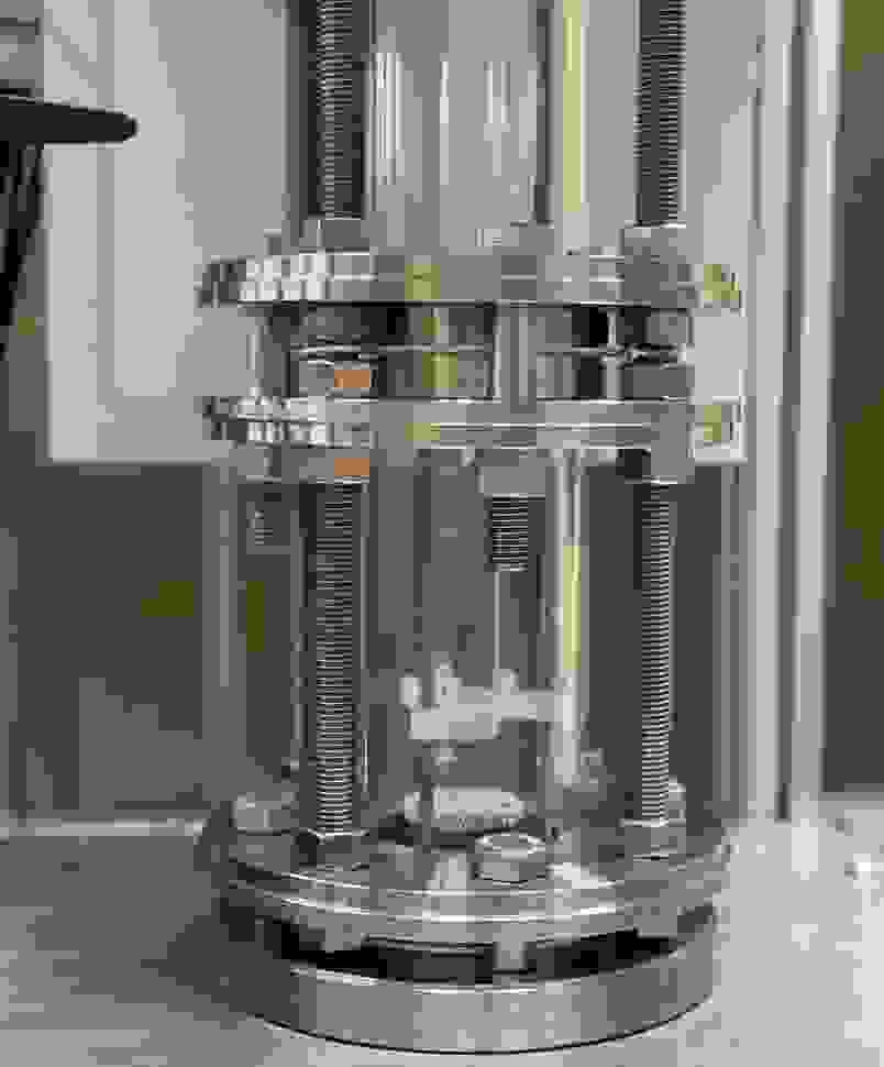 Detailansicht eines metallischen Druckbehälters mit einer transparenten Glassäule, die von Schrauben und Muttern fixiert wird.