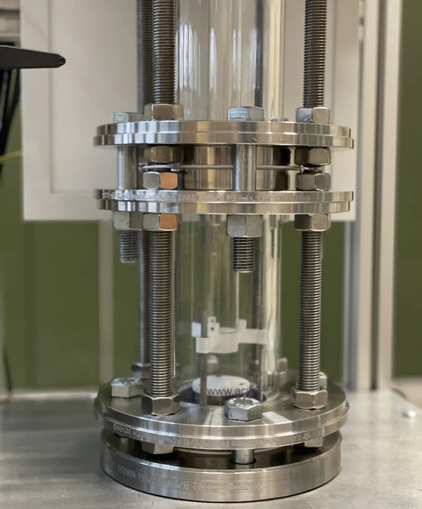 Detailansicht eines metallischen Druckbehälters mit einer transparenten Glassäule, die von Schrauben und Muttern fixiert wird.