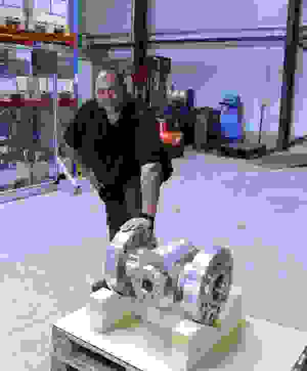 Ein Mann steht auf einem Werkstattboden neben einem großen, metallischen Rohrstück, das auf Holzklötzen liegt. Im Hintergrund sind Regale und Werkzeuge sichtbar.
