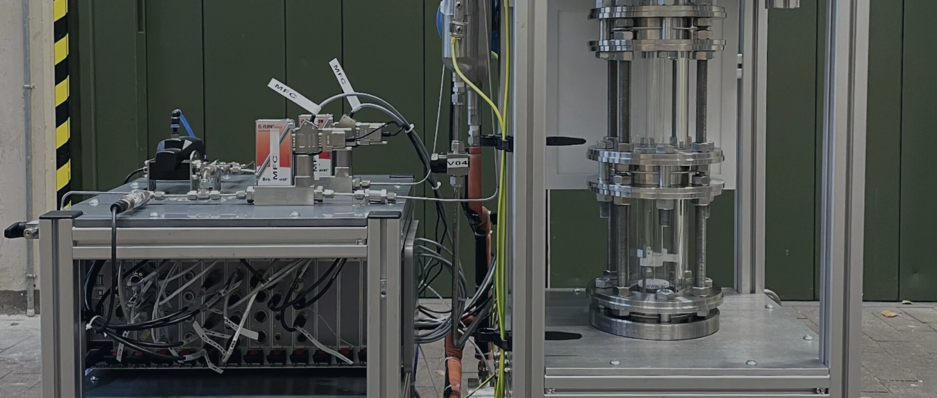 A technical device with two main parts: on the left, a control unit with pipes and sensors; on the right, a transparent pressure vessel system.