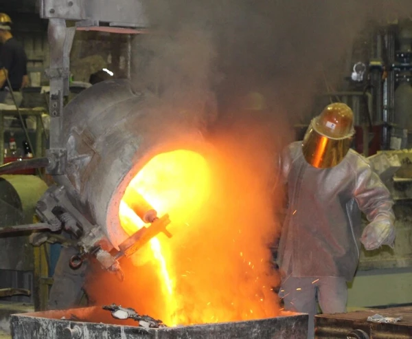 Ein Arbeiter in Schutzausrüstung gießt glühendes Metall aus einem Schutzbehälter in eine Form, während Rauch und Funken aufsteigen.