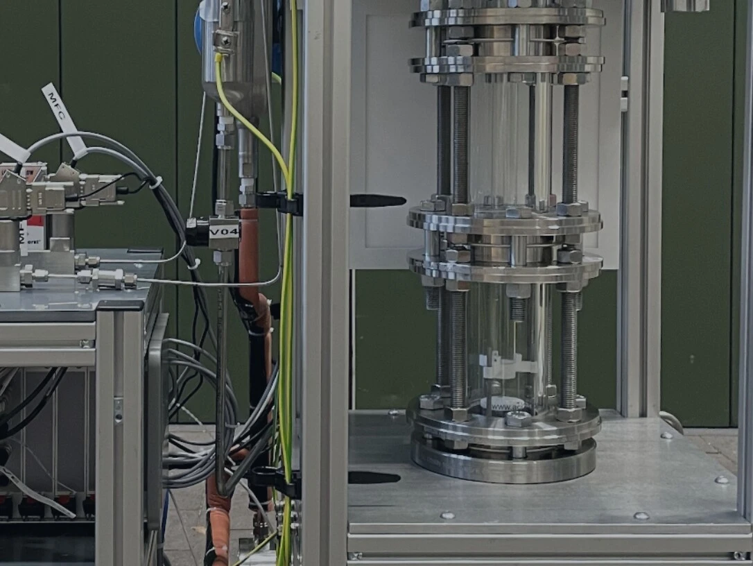 A technical device with two main parts: on the left, a control unit with pipes and sensors; on the right, a transparent pressure vessel system.