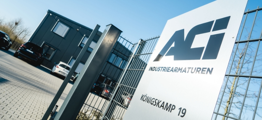 Un b&acirc;timent d'entreprise moderne avec une enseigne d'entr&eacute;e affichant la soci&eacute;t&eacute; 'ACI INDUSTRIEARMATUREN' &agrave; l'adresse 'K&Ouml;NIGSKAMP 19.' Des voitures gar&eacute;es sont visibles au premier plan.