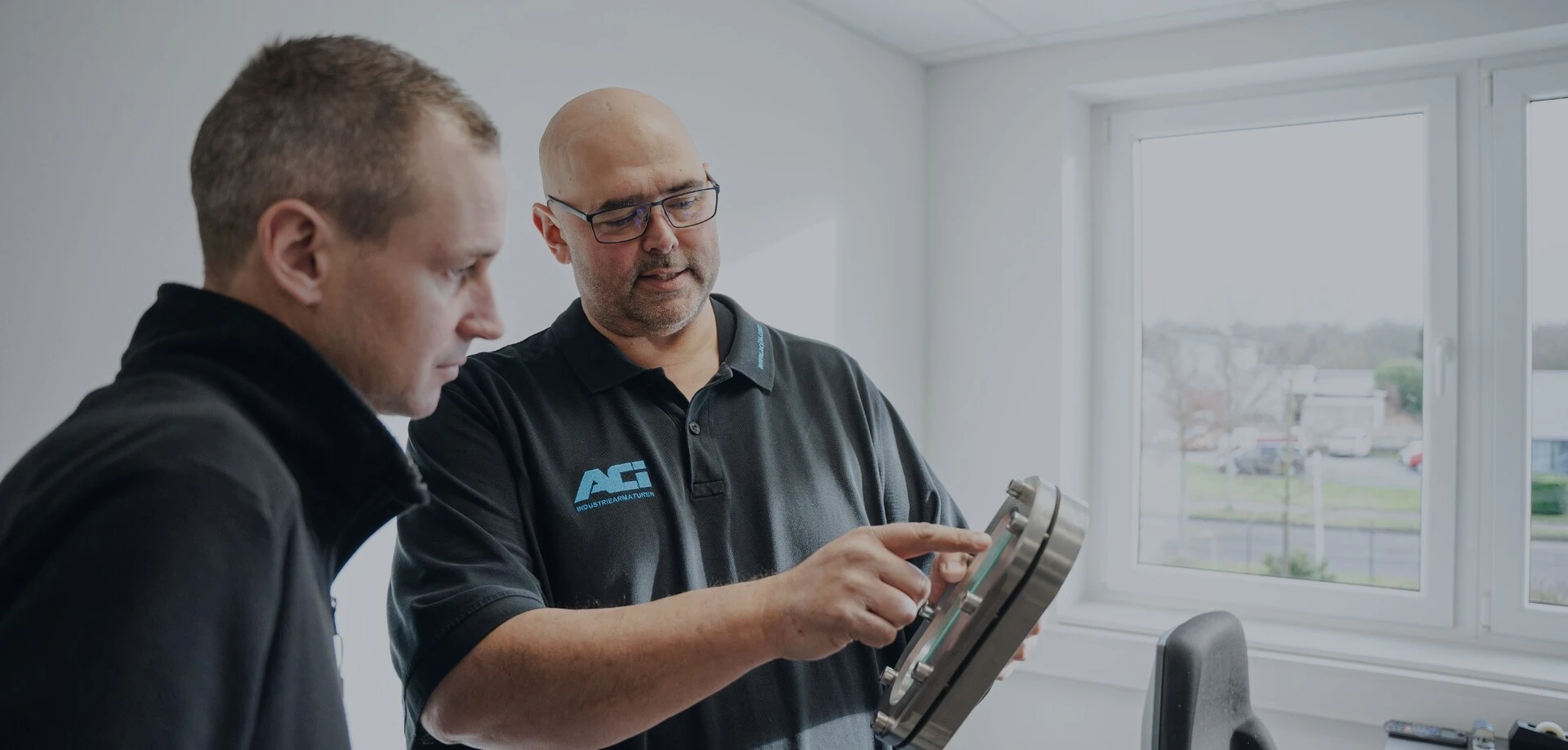 Twee mannen in gesprek over een industrieel onderdeel – een wijst met zijn vinger naar een rond metalen behuizing met kijkglas.