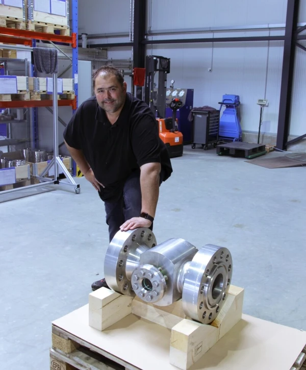 Een man staat op de vloer van een werkplaats naast een groot metalen pijpstuk dat op houten blokken ligt. Op de achtergrond zijn rekken en gereedschap zichtbaar.