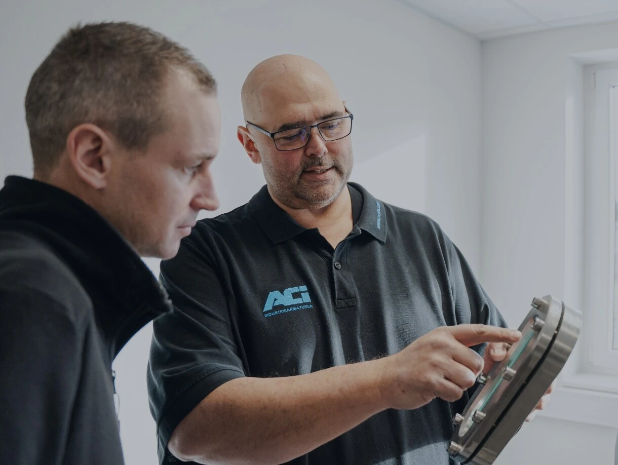 Twee mannen in gesprek over een industrieel onderdeel – een wijst met zijn vinger naar een rond metalen behuizing met kijkglas.