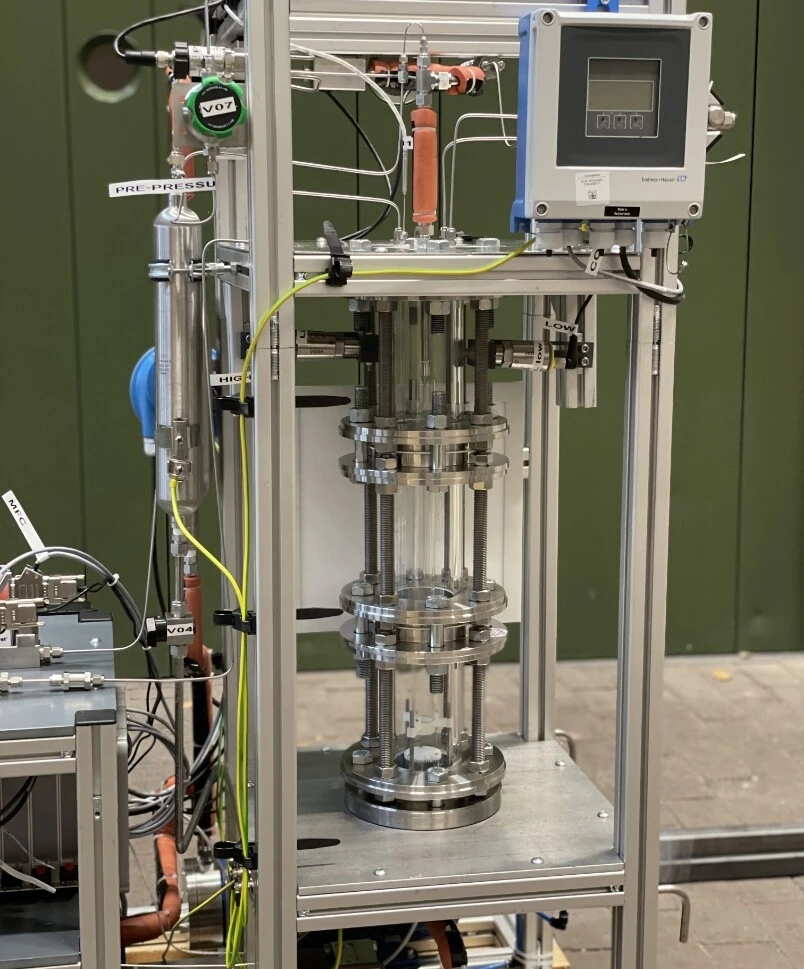 Metalowe urządzenie z przezroczystymi cylindrami i różnymi czujnikami, prawdopodobnie urządzenie doświadczalne lub stanowisko do testów ciśnieniowych.